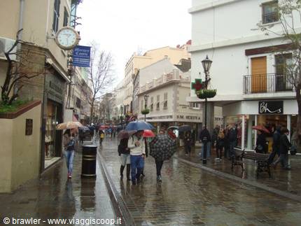 Shoppers with umbrellas on wet Main Street, Gibraltar, clock on white facade