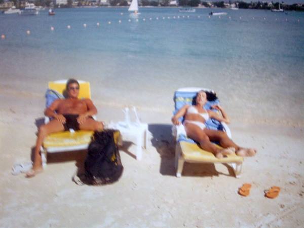 Man and woman in swimwear on yellow and blue loungers on sand, sea with boats