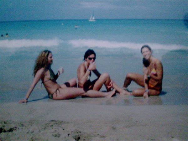 Three girls in swimsuits sitting on wet sand, sailboat on blue sea