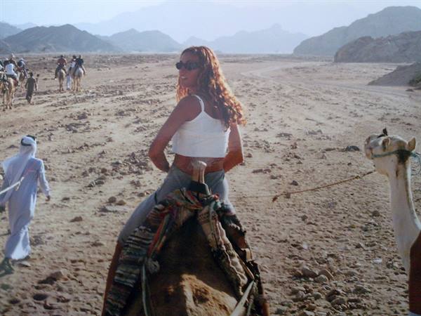 Tourist in white top riding a camel across rocky desert, guide and riders nearby