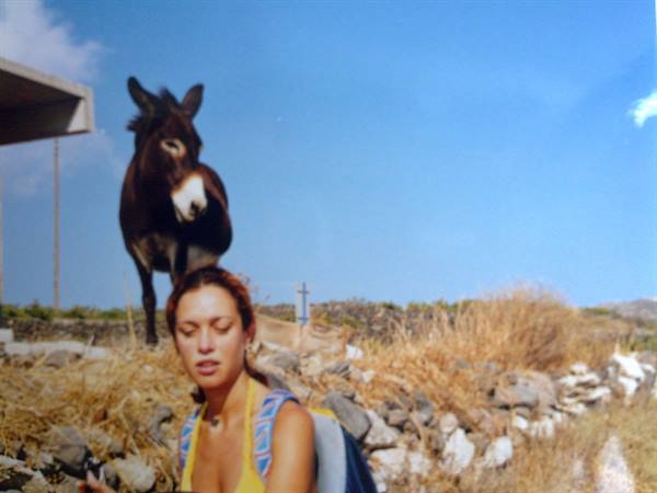 A blonde woman poses in front of a brown donkey among stone walls and dry grass