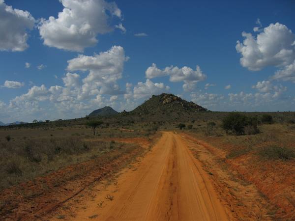 Red dirt road through savanna toward rocky hills, blue sky with clouds