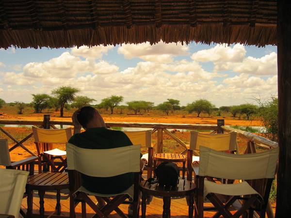 Man lounging in canvas chair under thatched roof, facing savanna with acacia trees