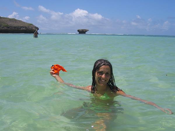 Girl in green swimsuit wades in turquoise water, raising an orange sarong near rocky cliffs