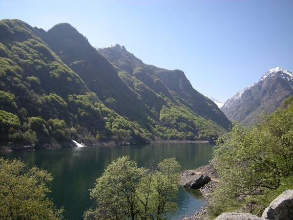 Dark green lake between forested mountains, small waterfall and snowy peaks under blue sky