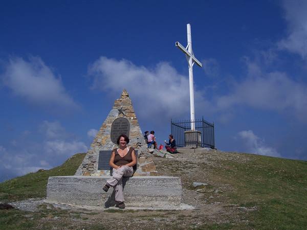 Woman sitting on stone memorial beside white cross on grassy summit