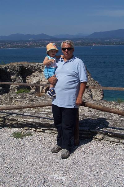 Grandfather holding toddler on Sirmione ruins, blue Lake Garda behind