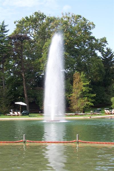 Tall fountain spraying water in a green park pond, sun loungers and umbrella under trees