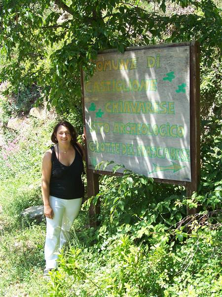 Woman beside Castiglione Chiavarese sign for the Màscarpe caves, amid greenery