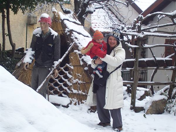 Woman holding child near snow-covered straw hut and scarecrow mannequin