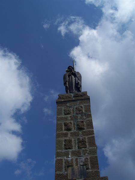 Stone statue of Padre Pio atop gray block pedestal against blue cloudy sky