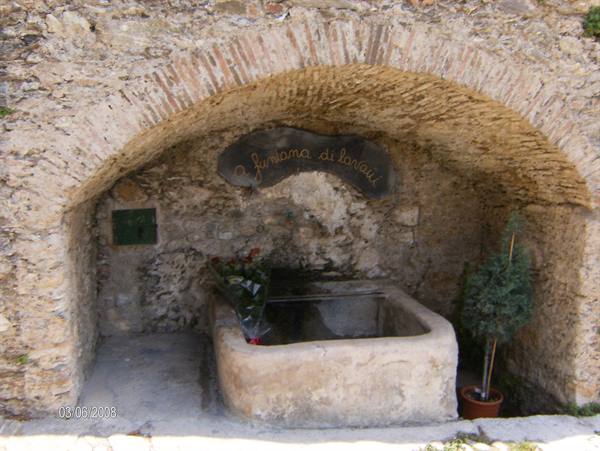 Old stone fountain under brick arch, painted sign above, potted plant beside