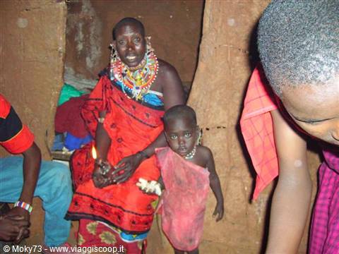 Maasai woman wearing beaded necklaces with a child inside a mud village hut