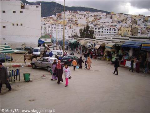 Women in djellabas cross the market of Moulay Idriss beneath white hillside houses