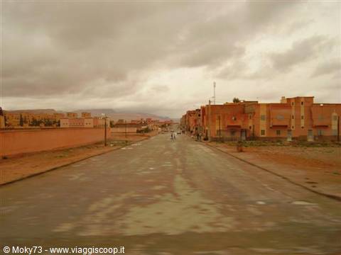 Wet road between pink earthen buildings under grey clouds, few pedestrians