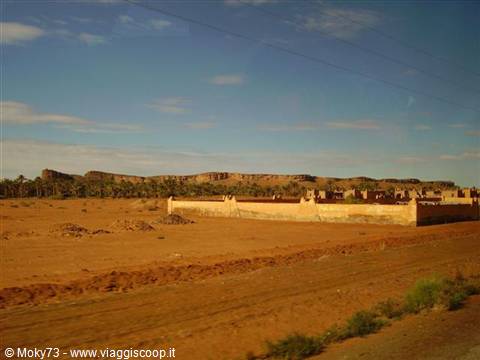 Ochre mud walls beside the road, palm grove and mountains under blue sky