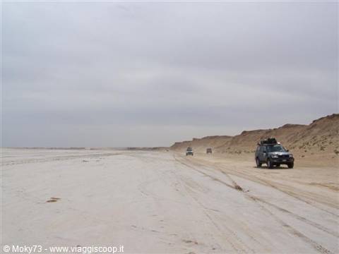 Loaded off-road vehicles cross the sandy flat under a grey sky
