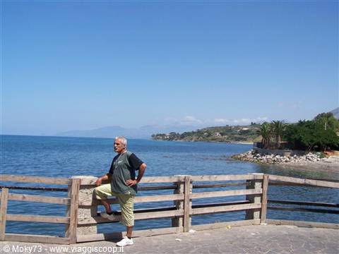 Man leaning on wooden railing, blue sea and Sicilian coastline in sunny weather