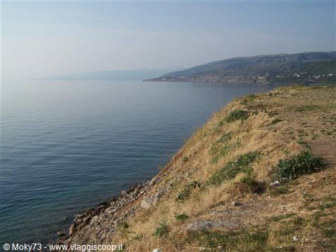 Rocky coast with dry grass above calm sea, hills in the haze