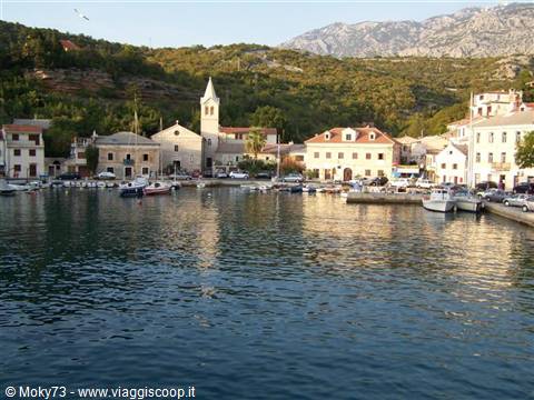 Boats moored in the harbor in front of colorful houses, white bell tower and green hills