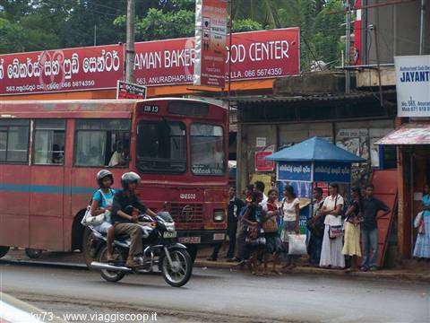 Red bus and motorbike passing a blue umbrella bus stop with waiting people