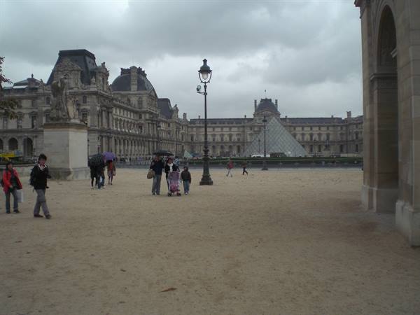 Tourists with umbrellas cross the Louvre's sandy courtyard toward the glass pyramid