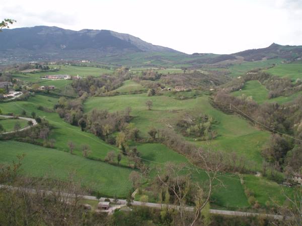 Green hills with fields, scattered houses and a road, mountains under cloudy sky