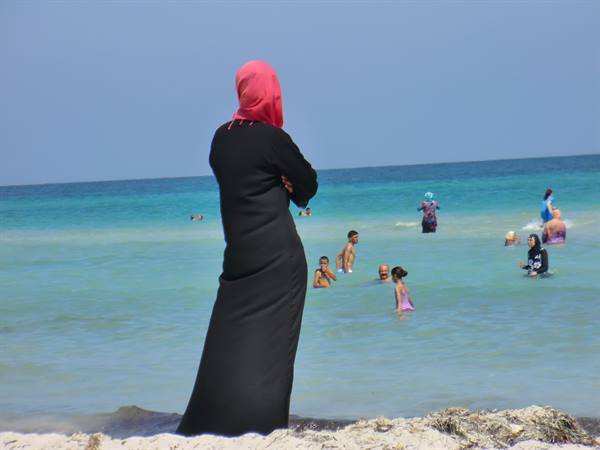 Woman in black abaya and pink headscarf watching swimmers in turquoise sea