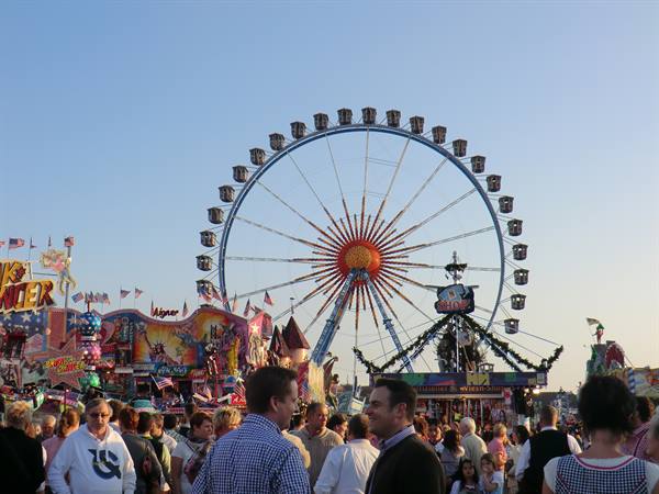 Fairground crowd, large blue Ferris wheel with gondolas behind colorful stalls