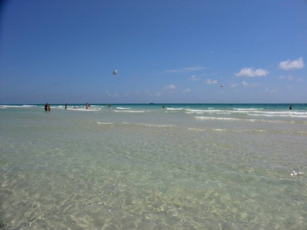 Bathers in shallow turquoise water on pale sand, blue sky with kites