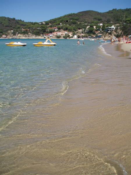 Sandy beach with yellow pedal boats on turquoise water, swimmers and green hills behind