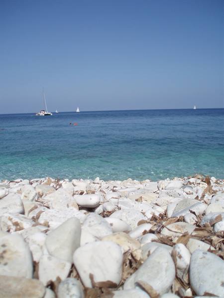White pebble beach, turquoise sea with sailboats under clear blue sky
