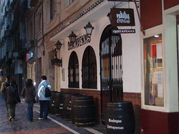 Bodega Quitapenas: pedestrians beside wooden barrels under the lit sign