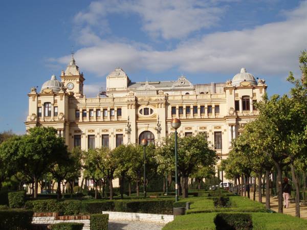 Málaga city hall, yellow facade with domes, surrounded by gardens under a cloudy sky