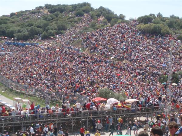 Huge crowd covering sunny green hills, spectators packed behind metal fences