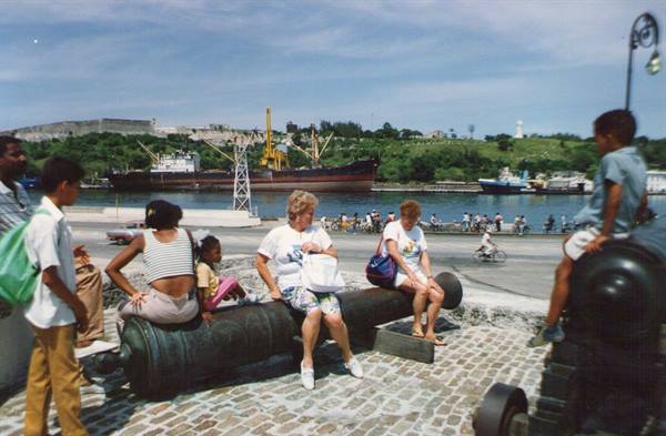 Tourists sitting on an old cannon, behind the bay with ships and El Morro fortress