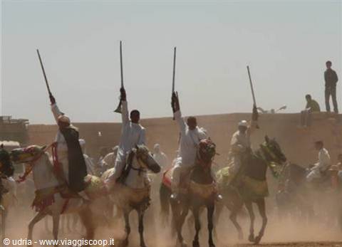 Horsemen in white raise rifles on decorated horses amid charging dust