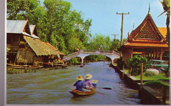 Woman in straw hat rows a boat on canal past wooden houses and white bridge