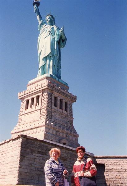 Two tourists beneath the green copper Statue of Liberty, blue sky