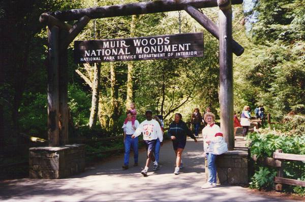 Visitors beneath the wooden entrance sign of Muir Woods National Monument, sunny forest