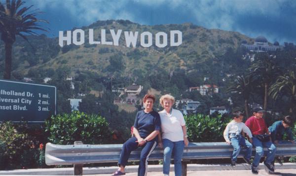 Two women sitting on guardrail, road sign, Hollywood sign on green hill
