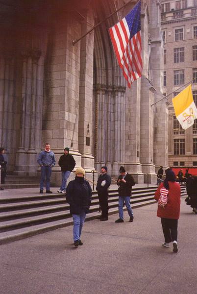 Tourists on stone steps outside St. Patrick's Cathedral under American and Vatican flags