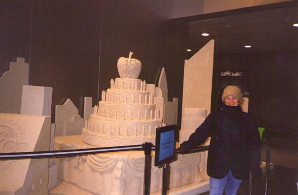 Woman in hat beside a sand-sculpted cake with an NYC apple topper