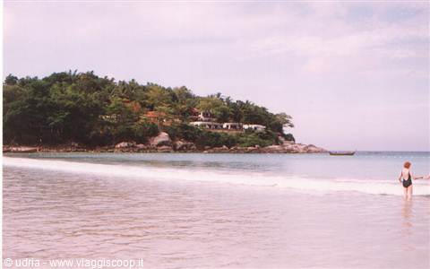 Woman in swimsuit walks in shallow water before a tropical hillside beach with boats