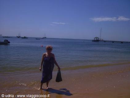 Woman with bag walking in shallow water on golden sand, boats and pier behind