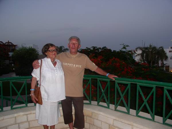 Couple posing on terrace with green railing, red flowers and palms behind