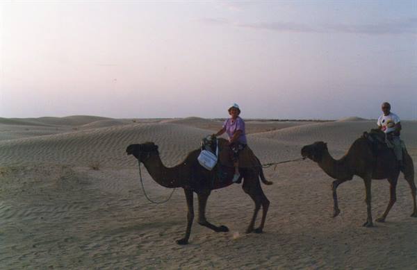 Two tourists ride camels across sandy dunes in soft evening light