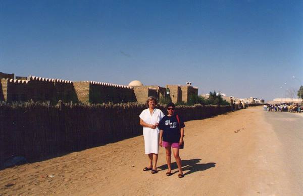 Two tourists on sandy road beside mud-brick houses and a dome