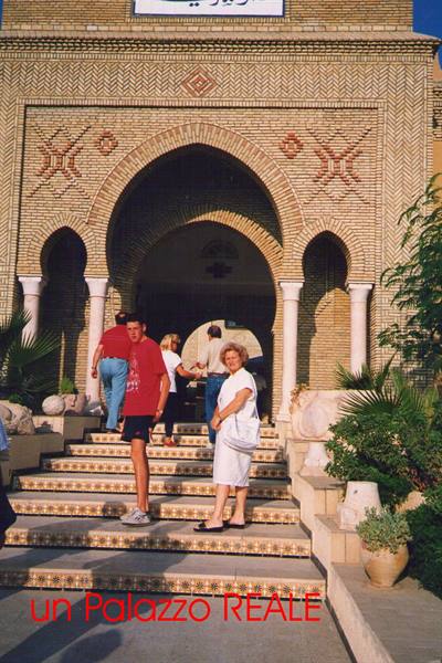 Woman in white and boy on steps of a brick palace with arches