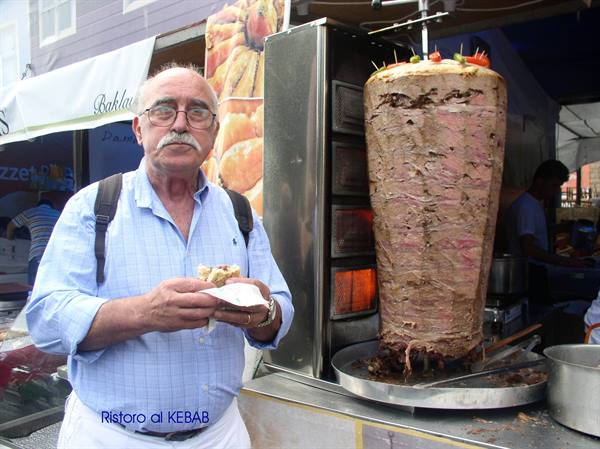 Man with backpack and glasses eats a sandwich beside the kebab spit